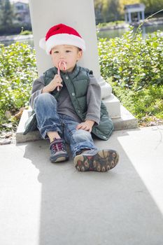 Cute Mixed Race Boy Sitting Wearing Christmas Santa Hat and Enjoying A Candy Cane.