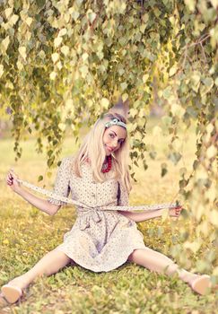 Beautiful woman smiling on grass in the park, sunny summer, happy people, outdoors. Pretty playful blonde girl like a doll in stylish dress, beads sitting under birch tree, enjoying nature.Toned,bokeh