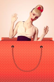 Fashion beauty woman in huge shopping bag surprised looks out of it on beige background, people, copyspace. Pinup funny blonde girl, hairstyle with fringe and pink bow. Shopaholic, vintage style