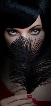 Fashion beauty portrait of sexy brunette woman with silky bob hairstyle on dark. Sensual lady mysteriously looks, covering face by black feather. Brown-eyed girl, makeup, red lips. People face closeup