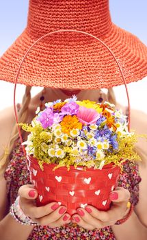 Woman with basket of wildflowers in hands. Young blonde boho girl in orange hat on summer meadow, people. Attractive joyfull, romantic style, floral sundress. Sunny day