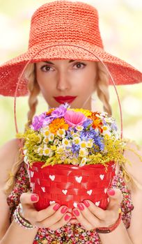 Woman with basket of wildflowers in hands. Young blonde boho girl in orange hat on summer meadow, people. Attractive joyfull, romantic style, floral sundress. Sunny day