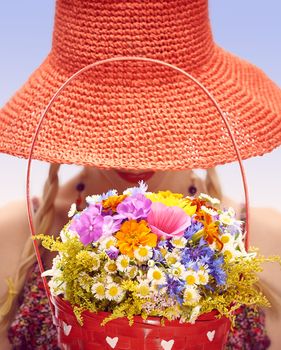 Young blonde boho woman on summer meadow with red basket of vivid wildflowers in hands. Attractive joyful girl with red lips in orange beach hat. Romantic style, floral sundress. Bokeh
