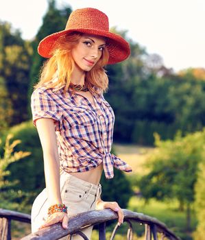 Beauty portrait of stylish playful smiling woman on wooden bridge in park, people, outdoors. Sunset, bokeh. Attractive happy girl in fashion clothes, hat enjoying nature in summer garden, lifestyle