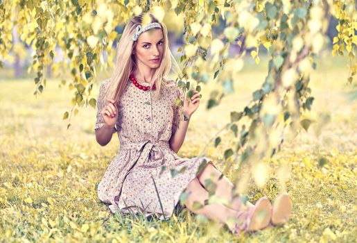 Beautiful woman dreaming on grass in the park, sunny summer, people, outdoors. Pretty playful blonde girl like a doll in stylish dress, beads sitting under birch tree, enjoying nature. Toned, bokeh