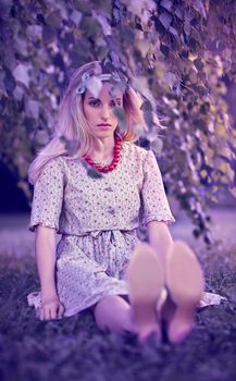 Beautiful woman on grass in the park, sadness, sunset summer, people, outdoors. Pretty playful blonde girl like a doll in stylish dress, beads sitting under birch tree, enjoying nature. Toned, bokeh
