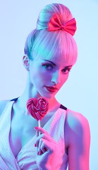 Fashion beauty portrait of sexy woman with heart shaped lollipop.  Sensual attractive playful blond girl. Luxury makeup, lips. Pin up hairstyle with fringe, pink bow. Colour creative toned, people