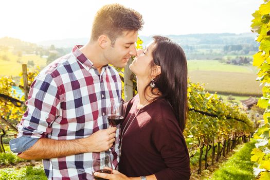 Photo of a young couple kissing and drinking wine in a vineyard.
