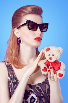 Beauty  fashion sexy slim model redhead woman in stylish mini dress holds loving teddy bear with red heart on blue. Sensual lady in sunglasses. Provocative young people, playful flirty mood. Copyspace
