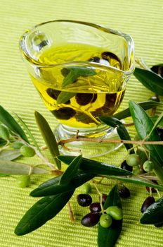 Olive Oil in Glass Gravy with Raw Green and Black Olives with Leafs closeup on Green Textile background