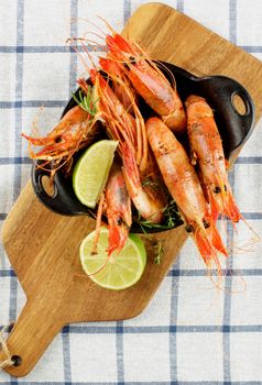 Delicious Roasted Shrimps in Black Cast Iron with Lime and Rosemary on Wooden Cutting Board closeup on Checkered Napkin background. Top View
