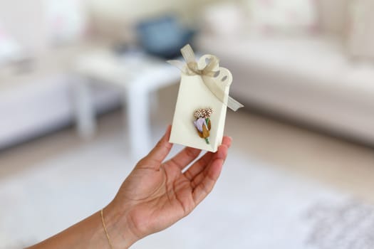 Close up detailed front view of hand-crafted white gift box holding on hand with small flowers and colorful ribbons.