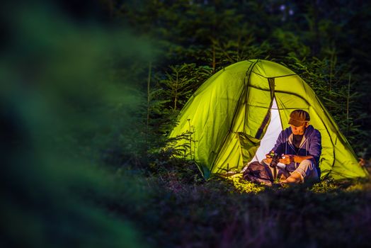 Overnight Forest Camping. Middle Age Caucasian Hiker with His Digital Camera and the Illuminated at Night Green Tent. Nighttime Mountains Hike and the Camping.