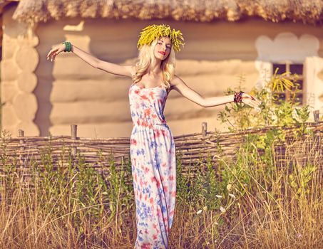 Boho Beauty woman in wreath enjoying nature, relax, harmony, outdoors, people, copyspace. Attractive boho girl in sundress near rustic house, summer meadow, flowers. Field, forest, ethnic background
