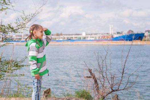 Girl in chilly weather stood by the river on which the floating ship and looks into the distance