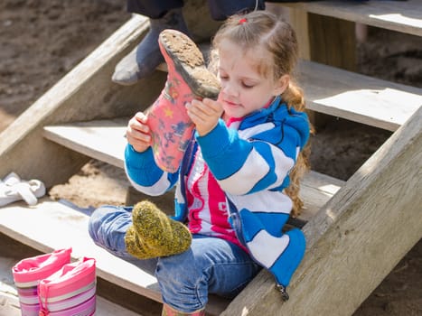 Four-year girl puts on boots sitting on the porch of a house in the village