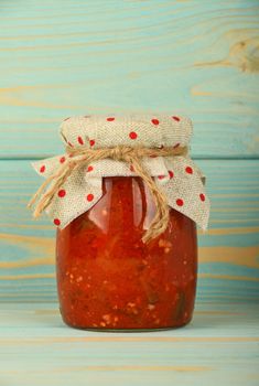 One glass jar of homemade pickled pepper, paprika and eggplant salad with dotted textile top decoration at blue painted vintage wooden surface