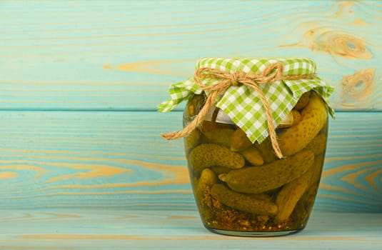 One glass jar of homemade pickled cucumbers with green checkered textile top decoration at blue painted vintage wooden surface