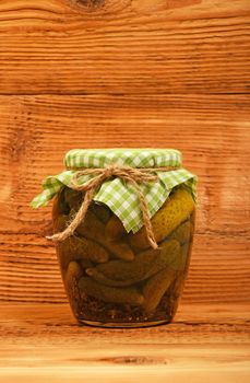 One glass jar of homemade pickled cucumbers with green checkered textile top decoration at brown painted vintage wooden surface