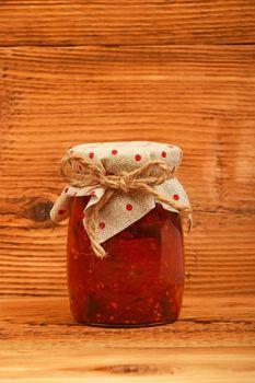 One glass jar of homemade pickled pepper, paprika and eggplant salad with dotted textile top decoration at brown vintage wooden surface