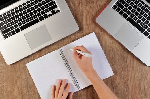 person writing in notebook infront of two laptops