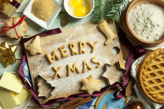 Christmas baking background with merry xmas made from cookies on a chopping board surrounded by cakes and recipe ingredients