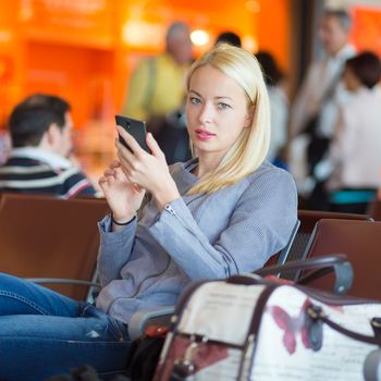 Casual blond young woman using her cell phone while waiting to board a plane at the departure gates. Wireless network hotspot enabling people to access internet conection. Public transport.