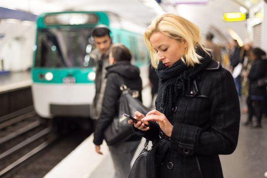 Young woman in winter coat with a cell phone in her hand waiting on the platform of a railway station for train to arrive. Public transport.  