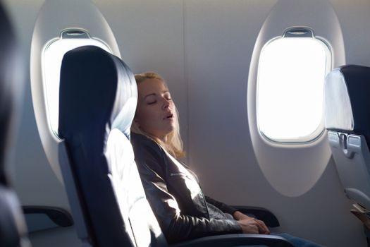 Tired blonde casual caucasian lady napping on uncomfortable seat while traveling by airplane. Commercial transportation by planes.