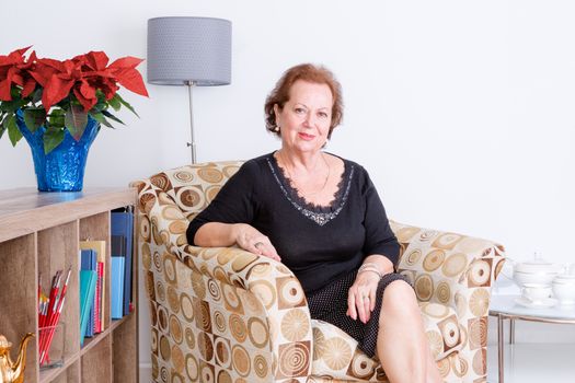 Gracious elegant senior woman relaxing at home in a comfortable armchair sitting smiling at the camera, festive red poinsettia flowers in a vase