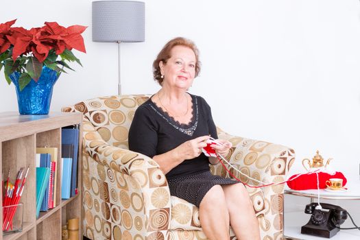 Stylish elderly lady sitting in an armchair at home relaxing and knitting with an old-fashioned rotary telephone and vase of festive red poinsettias alongside her