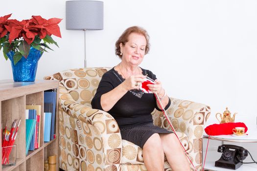 Happy senior lady enjoying her knitting as she sits at home in an armchair relaxing with a colorful arrangement of festive red poinsettia on a bookcase beside her