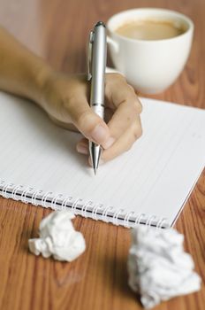 woman hand writing with pen on notebook.there are crumpled paper and coffee cup on wood table background