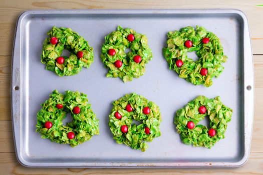 Creative colorful Christmas wreath cookies made with cornflakes and red candy on a metal oven tray, overhead closeup view