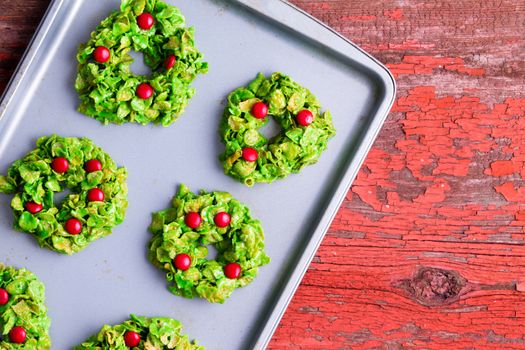 Freshly baked Christmas wreath cookies made with colorful green and red cornflakes and candy cooling on a metal baking tray on a red rustic table, overhead view with copy space
