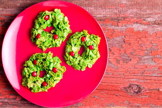 Festive colorful green Christmas cornflake wreath cookies displayed on a red plate on a cozy red rustic wooden table viewed from above with copy space