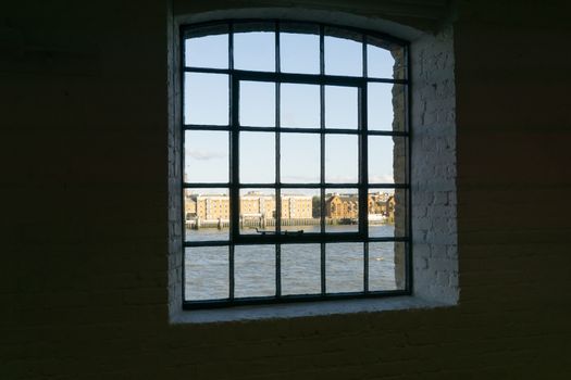 View framed by black surrounding wall to recent architectural buildings on other side River Thames through old wharf warehouse building windows from the dark interior at Wapping, London