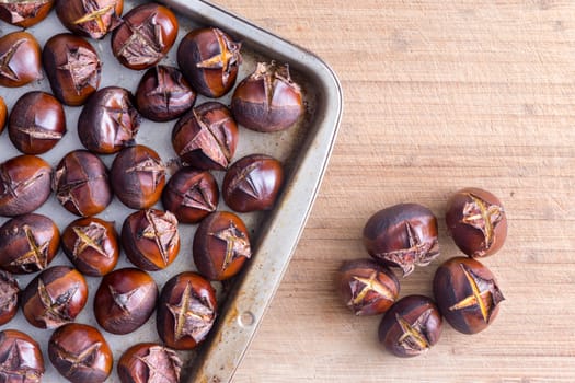 Metal tray of roasted chestnuts on wooden table next to five separate nuts