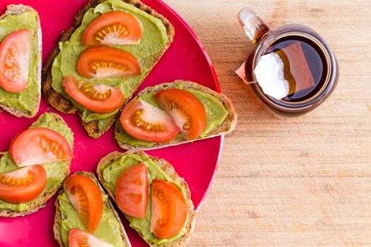Top view of avocado and tomato spread on bread on red plate with cup of herbal tea