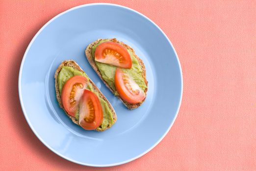 Top view of blue plate with healthy avocado and tomato sandwich over pink cloth background