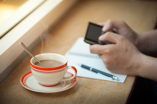 Male hand using smart phone on wood bar beside window with notebook and coffee cup in cafe in morning time