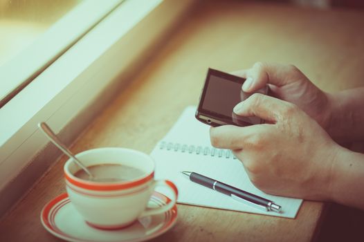 Male hand using smart phone on wood bar beside window with notebook and coffee cup in cafe in morning time with vintage filter effect