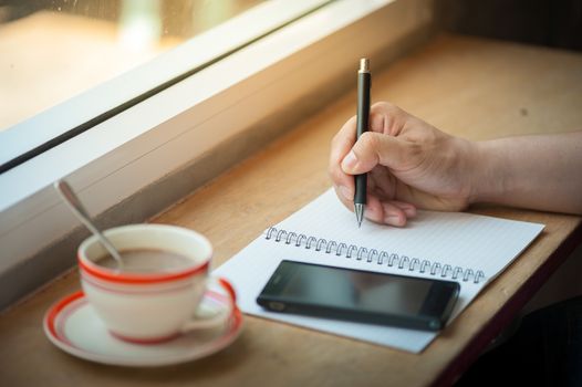 Male right hand writing on notebook with smart phone and coffee cup on wood bar beside window in cafe in morning time