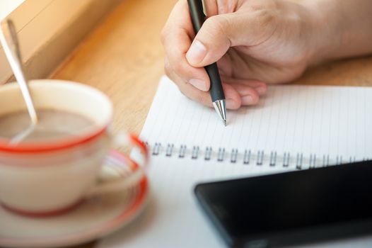 Male right hand writing on notebook with smart phone and coffee cup on wood bar beside window in cafe in morning time