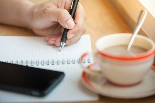 Male left hand writing on notebook with smart phone and coffee cup on wood bar beside window in cafe in morning time