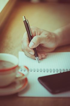 Male right hand writing on notebook with smart phone and coffee cup on wood bar beside window in cafe in morning time with vintage filter effect