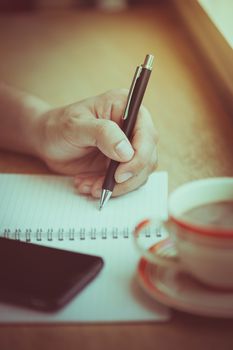 Male left hand writing on notebook with smart phone and coffee cup on wood bar beside window in cafe in morning time with vintage filter effect