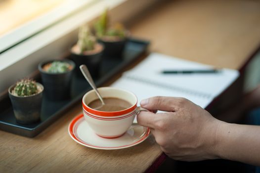 Male left hand holding coffee cup on wood bar beside window at cafe in morning time
