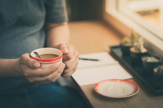 The man wearing grey t-shirt sitting beside window in cafe holding coffee cup in his hand in afternoon time with vintage filter effect