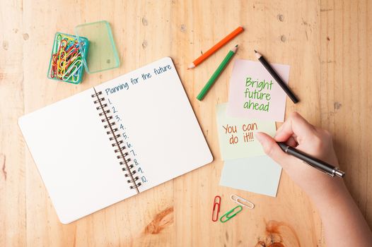 Female right hand writing "Bright future ahead you can do it" on notebook and sticky paper with paper clip, color pencil, and pen on wood table